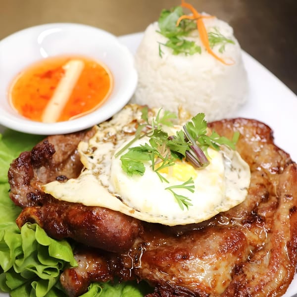 Grilled pork chop with a fried egg, rice, and sweet chili sauce on a white plate.