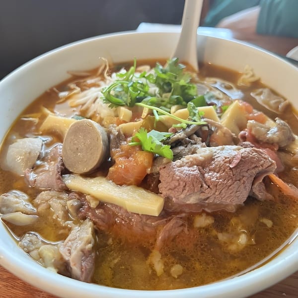 Close-up of a hearty Asian-fusion soup with beef, sausage, vegetables, and noodles in a white bowl.