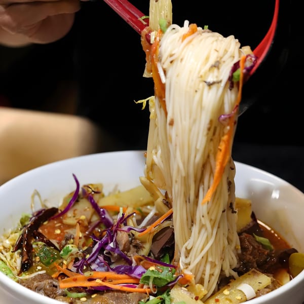 Red chopsticks lifting a large serving of noodles and vegetables from a bowl of B & K Asian Kitchen's Asian-fusion dish.