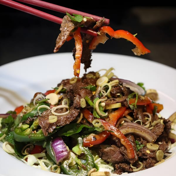 Chopsticks lifting a piece of beef and red pepper from a vibrant Asian-fusion salad.